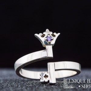 May include: A silver ring with a key design topped with a crown and a small purple gemstone. The ring is open and adjustable.