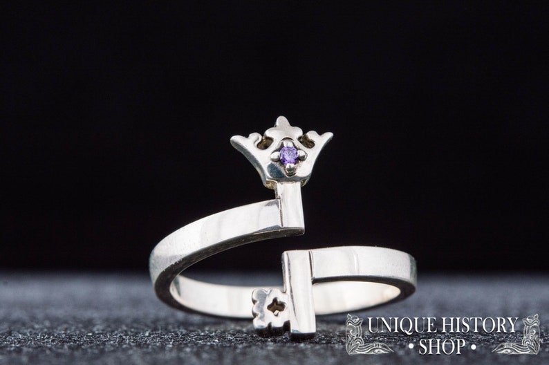 May include: A silver ring with a key design topped with a crown and a small purple gemstone. The ring is open and adjustable.