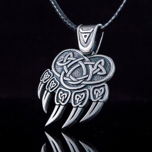 May include: Silver bear paw pendant necklace with a Celtic knot design. The pendant is attached to a black leather cord.