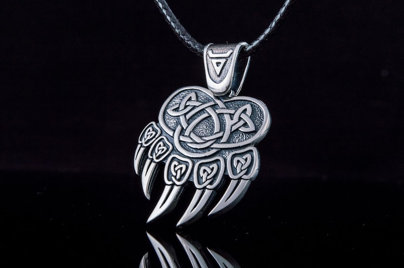 May include: Silver bear paw pendant necklace with a Celtic knot design. The pendant is attached to a black leather cord.