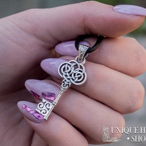 K&ouml;nnte beinhalten: Ein silberner Schl&uuml;sselanh&auml;nger mit einem keltischen Knotendesign. Der Anh&auml;nger h&auml;ngt an einer schwarzen Schnur und wird von einer Hand mit rosa Nagellack gehalten.