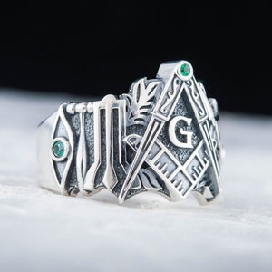 Könnte beinhalten: Ein silberner Ring mit Freimaurer-Symbolen, darunter Winkel und Zirkel mit dem Buchstaben "G". Der Ring hat zwei smaragdgrüne Edelsteine und detaillierte Gravuren.