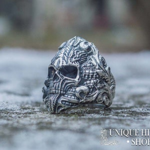 Peut inclure: Une bague en argent d&eacute;taill&eacute;e en forme de cr&acirc;ne avec des gravures complexes. La bague pr&eacute;sente un design de cr&acirc;ne avec des traits faciaux d&eacute;taill&eacute;s et des motifs orn&eacute;s. La bague est pos&eacute;e sur un fond flou et le texte "UNIQUE HISTORY SHOP" est visible.