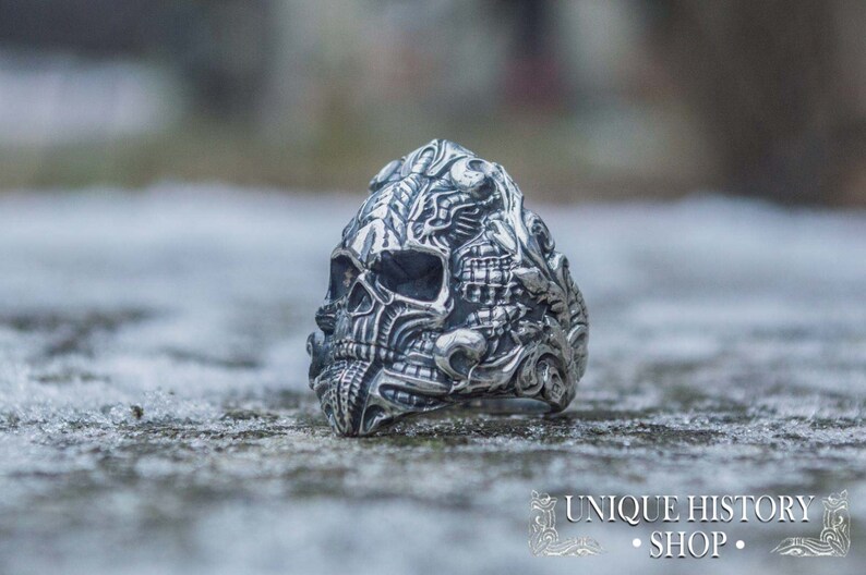 Peut inclure: Une bague en argent d&eacute;taill&eacute;e en forme de cr&acirc;ne avec des gravures complexes. La bague pr&eacute;sente un design de cr&acirc;ne avec des traits faciaux d&eacute;taill&eacute;s et des motifs orn&eacute;s. La bague est pos&eacute;e sur un fond flou et le texte "UNIQUE HISTORY SHOP" est visible.