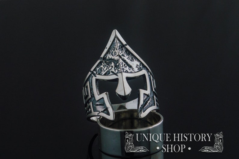Pode incluir: Um anel de prata com um design detalhado de um capacete espartano. O anel est&aacute; sobre um fundo preto e o texto "UNIQUE HISTORY SHOP" &eacute; vis&iacute;vel abaixo do anel.