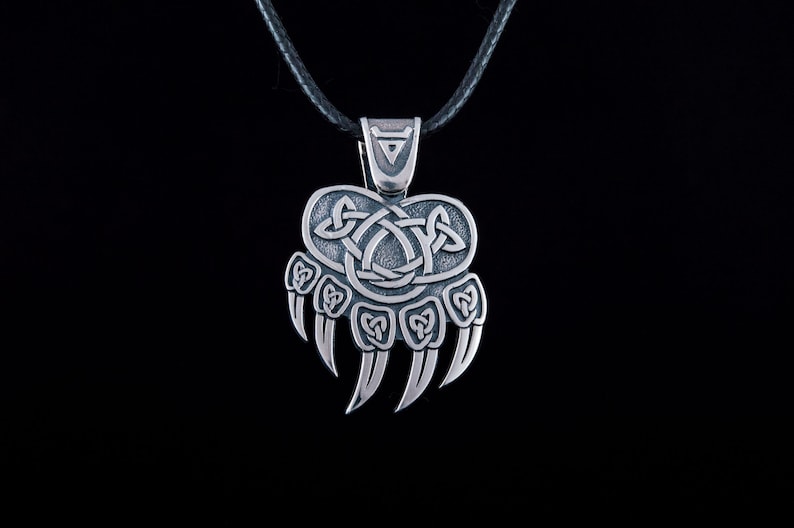 May include: Silver bear paw pendant necklace with a Celtic knot design. The pendant is on a black leather cord.