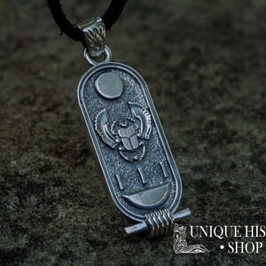 May include: Silver pendant featuring a scarab beetle with wings, surrounded by hieroglyphs and a crescent moon. The pendant is attached to a black cord.