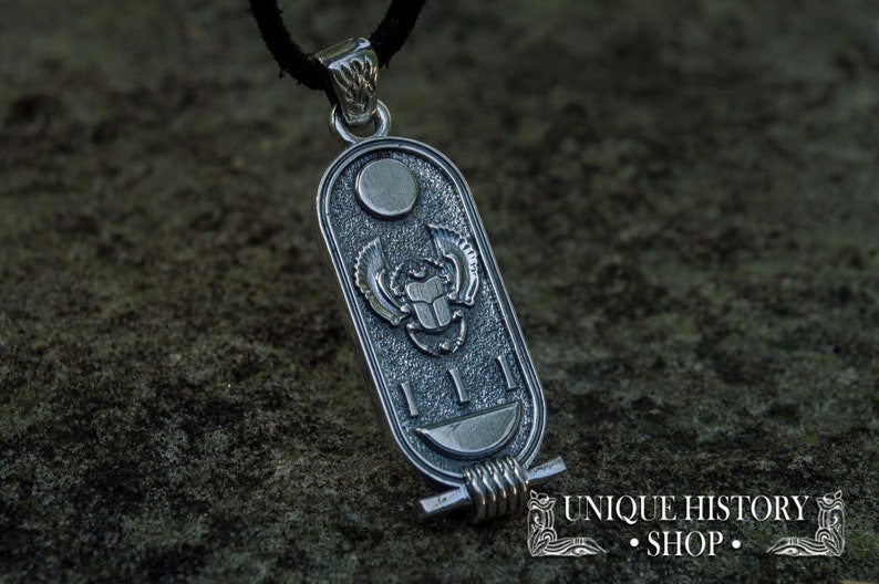 May include: Silver pendant featuring a scarab beetle with wings, surrounded by hieroglyphs and a crescent moon. The pendant is attached to a black cord.