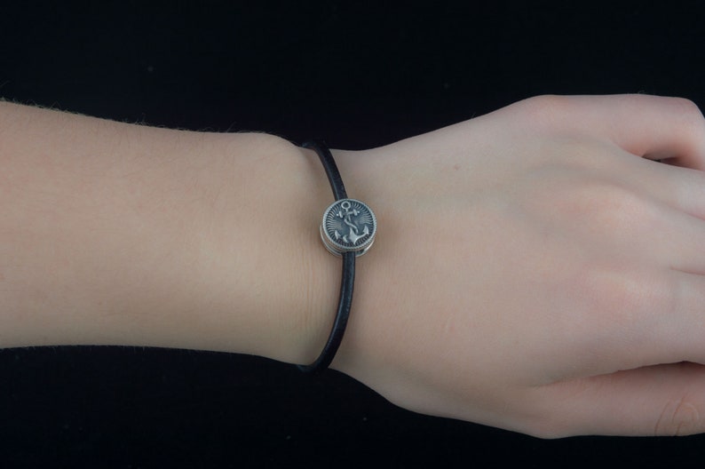 Puede incluir: Un brazalete de cuero negro con un dije de plata con dise&ntilde;o de ancla.