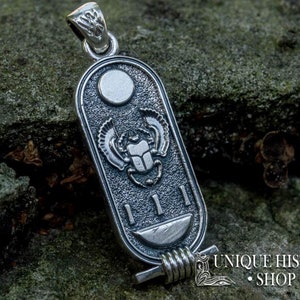 May include: Silver pendant featuring a scarab beetle with wings, a sun disc, and hieroglyphs. The pendant is on a textured, green surface. "UNIQUE HISTORY SHOP" is visible in the bottom right corner.