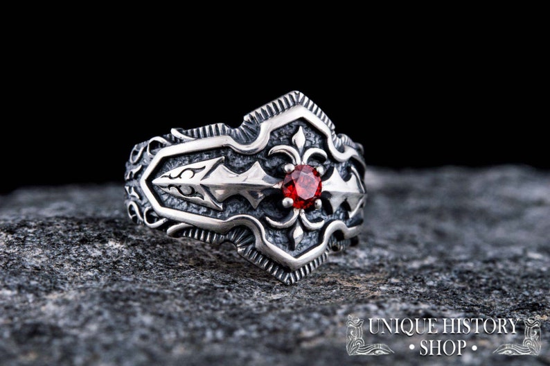 May include: A silver-toned ring with an ornate design featuring a central red gemstone. The ring has a detailed, textured pattern with a fleur-de-lis motif and arrow-like elements. The shop's name is visible at the bottom.
