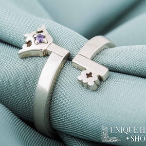 May include: A silver ring with a key design featuring a purple gemstone. The ring is on a light blue fabric background.