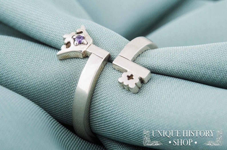 May include: A silver ring with a key design featuring a purple gemstone. The ring is on a light blue fabric background.