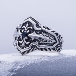 May include: A silver-colored ring with an ornate design. The ring features a central black gemstone and detailed patterns. The design includes floral and arrow-like motifs, creating a vintage or gothic aesthetic. The ring is displayed on a textured surface.