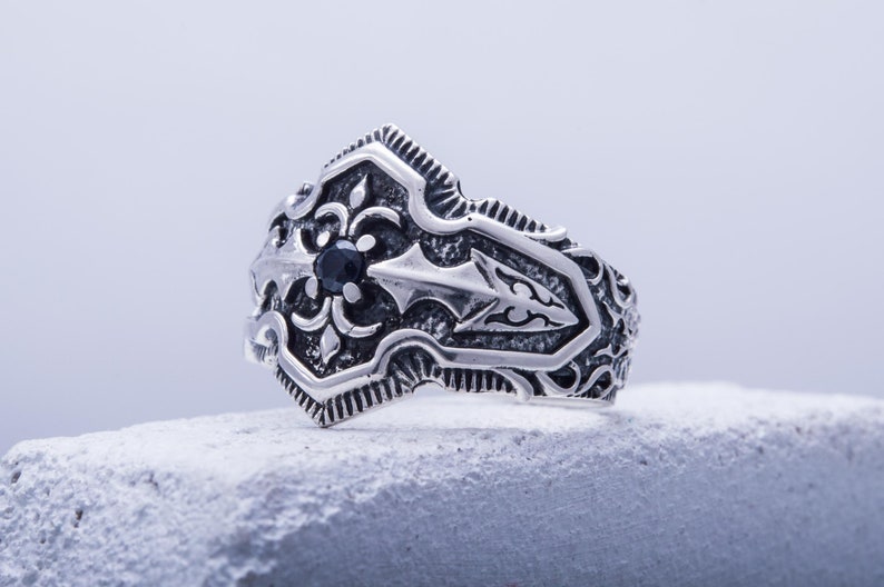 May include: A silver-colored ring with an ornate design. The ring features a central black gemstone and detailed patterns. The design includes floral and arrow-like motifs, creating a vintage or gothic aesthetic. The ring is displayed on a textured surface.