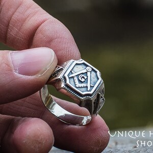 K&ouml;nnte beinhalten: Ein silberner Ring mit einem detaillierten Design, das einen Kompass und ein Geodreieck zeigt, ein h&auml;ufiges Symbol der Freimaurerei.