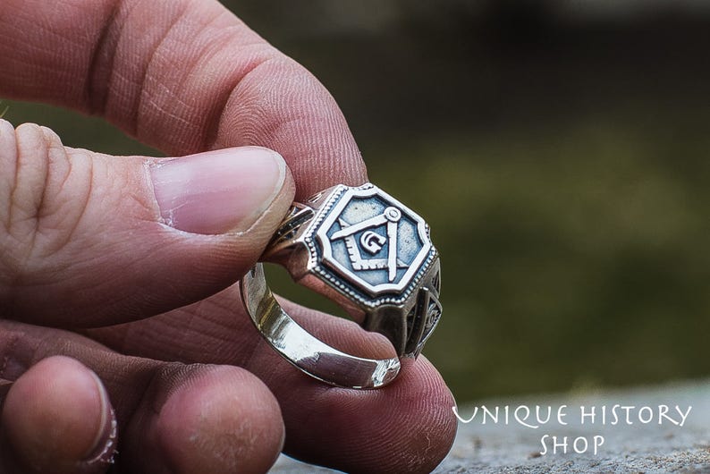 K&ouml;nnte beinhalten: Ein silberner Ring mit einem detaillierten Design, das einen Kompass und ein Geodreieck zeigt, ein h&auml;ufiges Symbol der Freimaurerei.