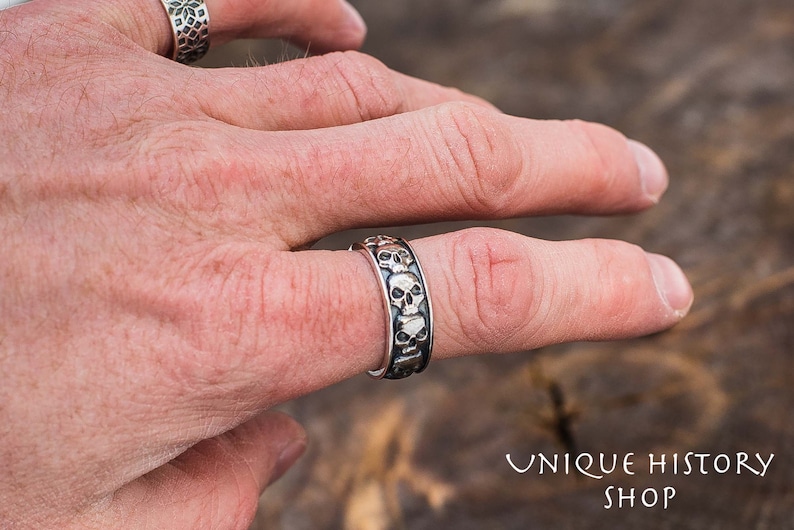 May include: A silver ring with a band of skulls. The ring is worn on a person's finger.