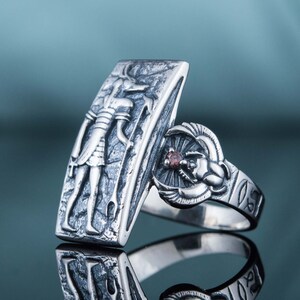 May include: A silver ring with an engraved image of an Egyptian god and a scarab beetle with a red stone. The ring is on a blue background.
