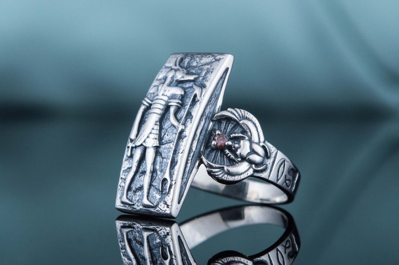 May include: A silver ring with an engraved image of an Egyptian god and a scarab beetle with a red stone. The ring is on a blue background.