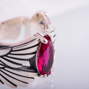 May include: A silver ring featuring a wing design and a large, teardrop-shaped, red gemstone. The ring has detailed feather-like engravings. The background is white.
