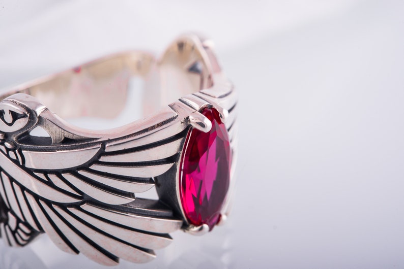 May include: A silver ring featuring a wing design and a large, teardrop-shaped, red gemstone. The ring has detailed feather-like engravings. The background is white.