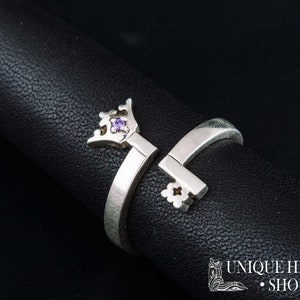 May include: A silver ring shaped like a key with a purple gemstone in the crown. The ring is open and can be adjusted to fit different finger sizes.