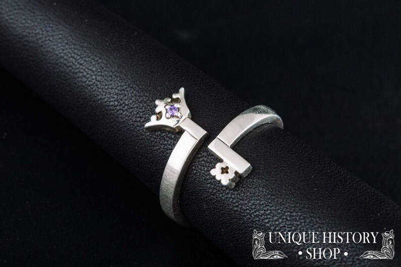 May include: A silver ring shaped like a key with a purple gemstone in the crown. The ring is open and can be adjusted to fit different finger sizes.