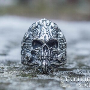 Peut inclure: Une bague argent&eacute;e d&eacute;taill&eacute;e en forme de cr&acirc;ne avec des gravures complexes. La bague pr&eacute;sente un design de cr&acirc;ne avec des traits faciaux d&eacute;taill&eacute;s et des ornements. La bague est pos&eacute;e sur un fond flou et enneig&eacute;. Le texte "UNIQUE HISTORY SHOP" est visible.