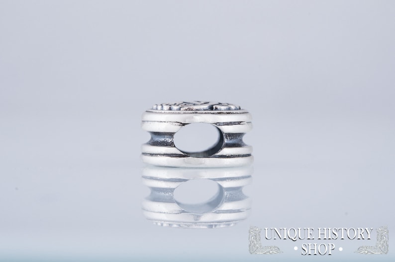 Puede incluir: Un charm de plata con un dise&ntilde;o detallado y un agujero en el centro. El charm tiene forma cil&iacute;ndrica con la parte superior e inferior redondeadas.