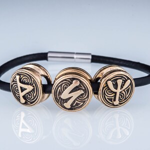 May include: A black leather bracelet with three gold-toned metal beads. Each bead features a different Norse rune symbol.