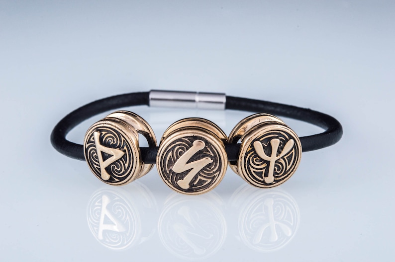 May include: A black leather bracelet with three gold-toned metal beads. Each bead features a different Norse rune symbol.