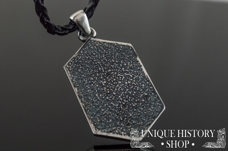 May include: Silver pendant with a textured, octagonal shape. The pendant is attached to a black leather cord.