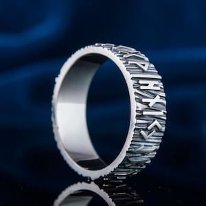 Op de afbeelding: Een zilveren ring met een getextureerde band en runen gegraveerd rond de buitenkant. De runen zijn verticaal geori&euml;nteerd.