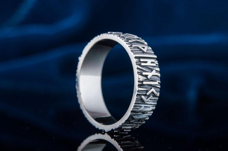 Op de afbeelding: Een zilveren ring met een getextureerde band en runen gegraveerd rond de buitenkant. De runen zijn verticaal geori&euml;nteerd.