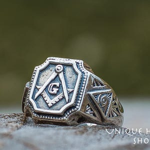 K&ouml;nnte beinhalten: Ein silberner Freimaurer-Ring mit einem Quadrat- und Zirkel-Design und dem Buchstaben "G" in der Mitte. Der Ring hat auch ein dreieckiges Design mit einem Auge in der Mitte.