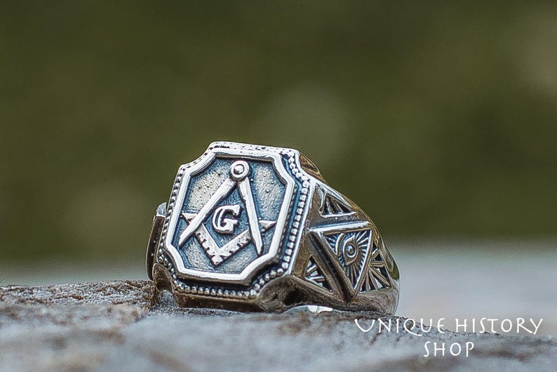 K&ouml;nnte beinhalten: Ein silberner Freimaurer-Ring mit einem Quadrat- und Zirkel-Design und dem Buchstaben "G" in der Mitte. Der Ring hat auch ein dreieckiges Design mit einem Auge in der Mitte.
