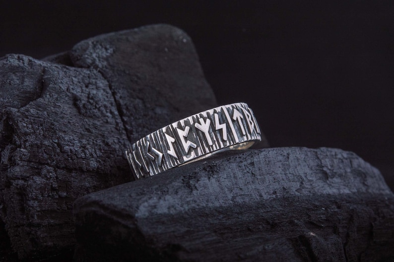 Op de afbeelding: Een zilveren ring met een getextureerd hout-achtig ontwerp en gegraveerde Noorse runen op de band.
