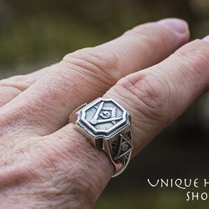 K&ouml;nnte beinhalten: Ein silberner Ring mit einem freimaurerischen Symbol, bestehend aus einem Zirkel und einem Winkelma&szlig;, die ein allsehendes Auge umgeben. Der Ring wird an einem Finger getragen.