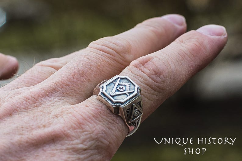 K&ouml;nnte beinhalten: Ein silberner Ring mit einem freimaurerischen Symbol, bestehend aus einem Zirkel und einem Winkelma&szlig;, die ein allsehendes Auge umgeben. Der Ring wird an einem Finger getragen.