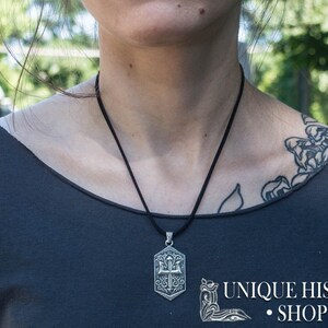 May include: A silver pendant necklace with a detailed design of a trident and other symbols. The necklace is on a black cord and is being worn by a person with a tattoo on their shoulder.