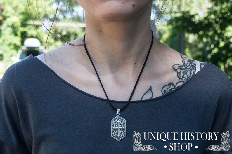 May include: A silver pendant necklace with a detailed design of a trident and other symbols. The necklace is on a black cord and is being worn by a person with a tattoo on their shoulder.