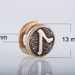 May include: A gold-colored metal charm with a black rune symbol on a circular surface. The rune is surrounded by a swirling pattern. The charm is 13 mm in diameter and 8 mm thick.