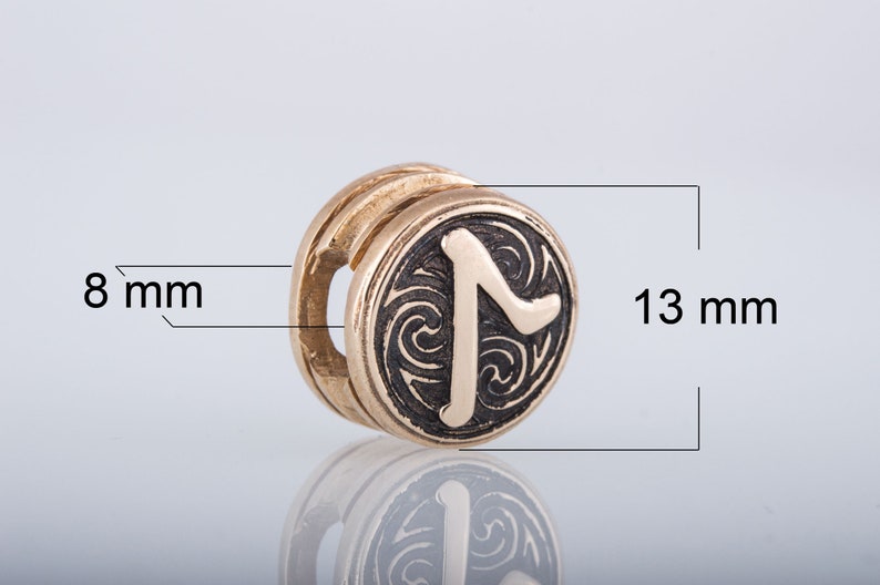 May include: A gold-colored metal charm with a black rune symbol on a circular surface. The rune is surrounded by a swirling pattern. The charm is 13 mm in diameter and 8 mm thick.