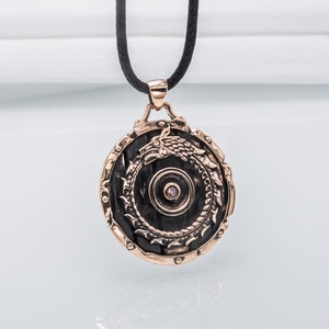 Pode incluir: Um pingente redondo de bronze com um design detalhado de um drag&atilde;o envolto em torno de um c&iacute;rculo. O pingente tem uma pequena gema rosa no centro.