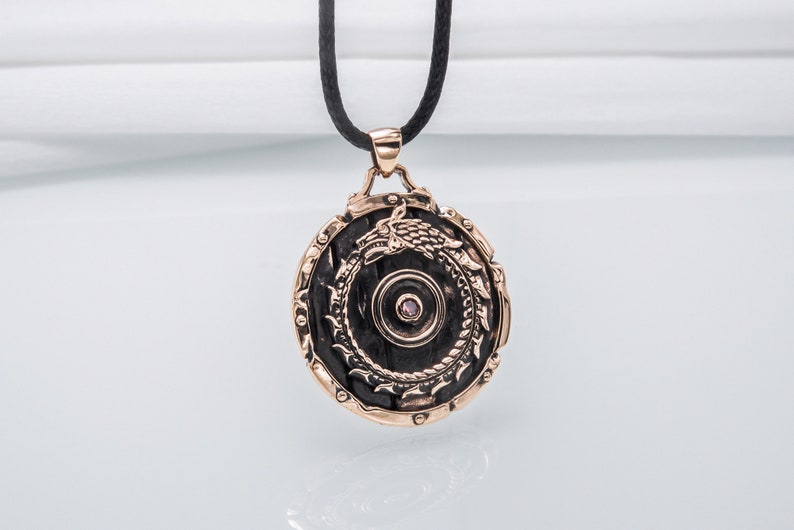 Pode incluir: Um pingente redondo de bronze com um design detalhado de um drag&atilde;o envolto em torno de um c&iacute;rculo. O pingente tem uma pequena gema rosa no centro.