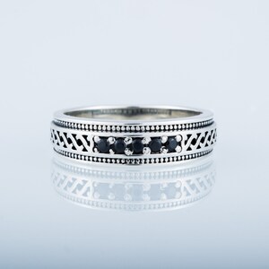 May include: A silver band ring with a black stone inlay and a decorative pattern. The ring is on a white background.