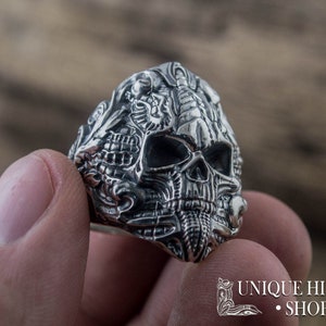 Peut inclure: Une bague en argent d&eacute;taill&eacute;e repr&eacute;sentant un cr&acirc;ne avec des gravures complexes. La bague pr&eacute;sente un design de cr&acirc;ne avec des dents et des orbites visibles. La bague est tenue dans une main, mettant en valeur le savoir-faire. La bague est de Unique History Shop.