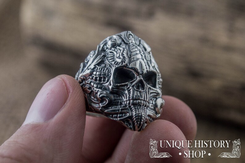 Peut inclure: Une bague en argent d&eacute;taill&eacute;e repr&eacute;sentant un cr&acirc;ne avec des gravures complexes. La bague pr&eacute;sente un design de cr&acirc;ne avec des dents et des orbites visibles. La bague est tenue dans une main, mettant en valeur le savoir-faire. La bague est de Unique History Shop.