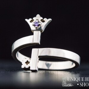 May include: A silver ring shaped like a key with a small purple gemstone on the crown. The ring is open and adjustable.